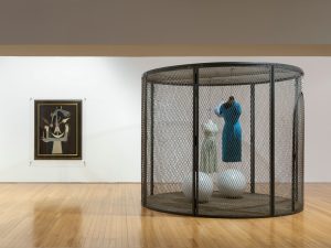 exposição de arte contemporânea apresentando manequins dentro de uma estrutura de rede, com uma pintura ao fundo.