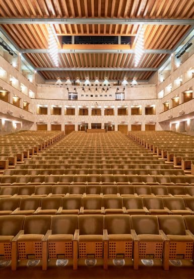 Auditório amplo e moderno com assentos de couro bege, iluminação sofisticada e teto de madeira, ideal para eventos culturais e apresentações musicais.