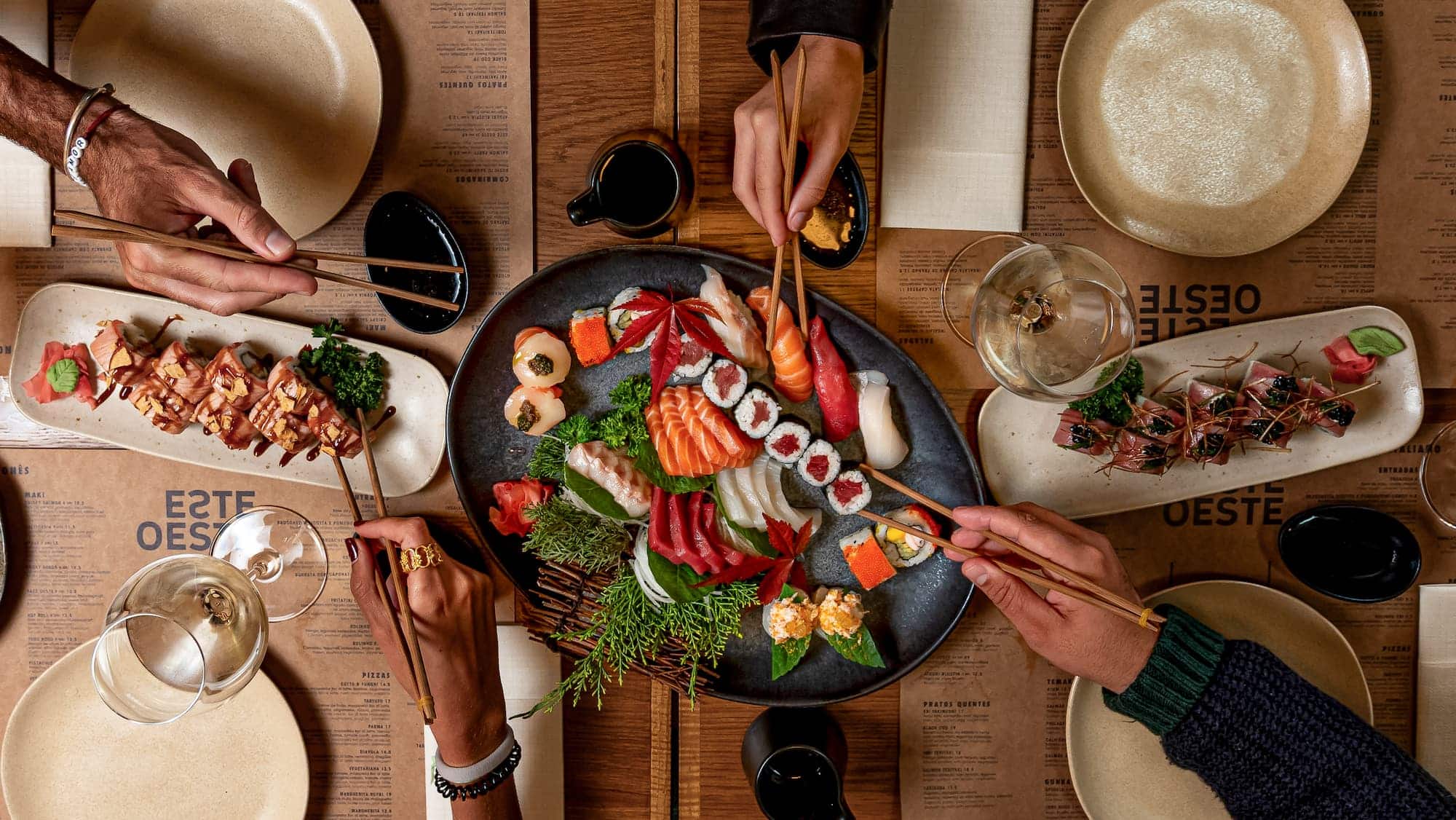 http://Pessoas%20comendo%20sushi%20em%20uma%20mesa%20de%20jantar%20com%20pratos,%20copos%20e%20um%20comedouro%20de%20sushi%20na%20imagem,%20criando%20uma%20experiência%20gastronômica%20de%20culinária%20japonesa,%20no%20restaurante%20EsteOeste.
