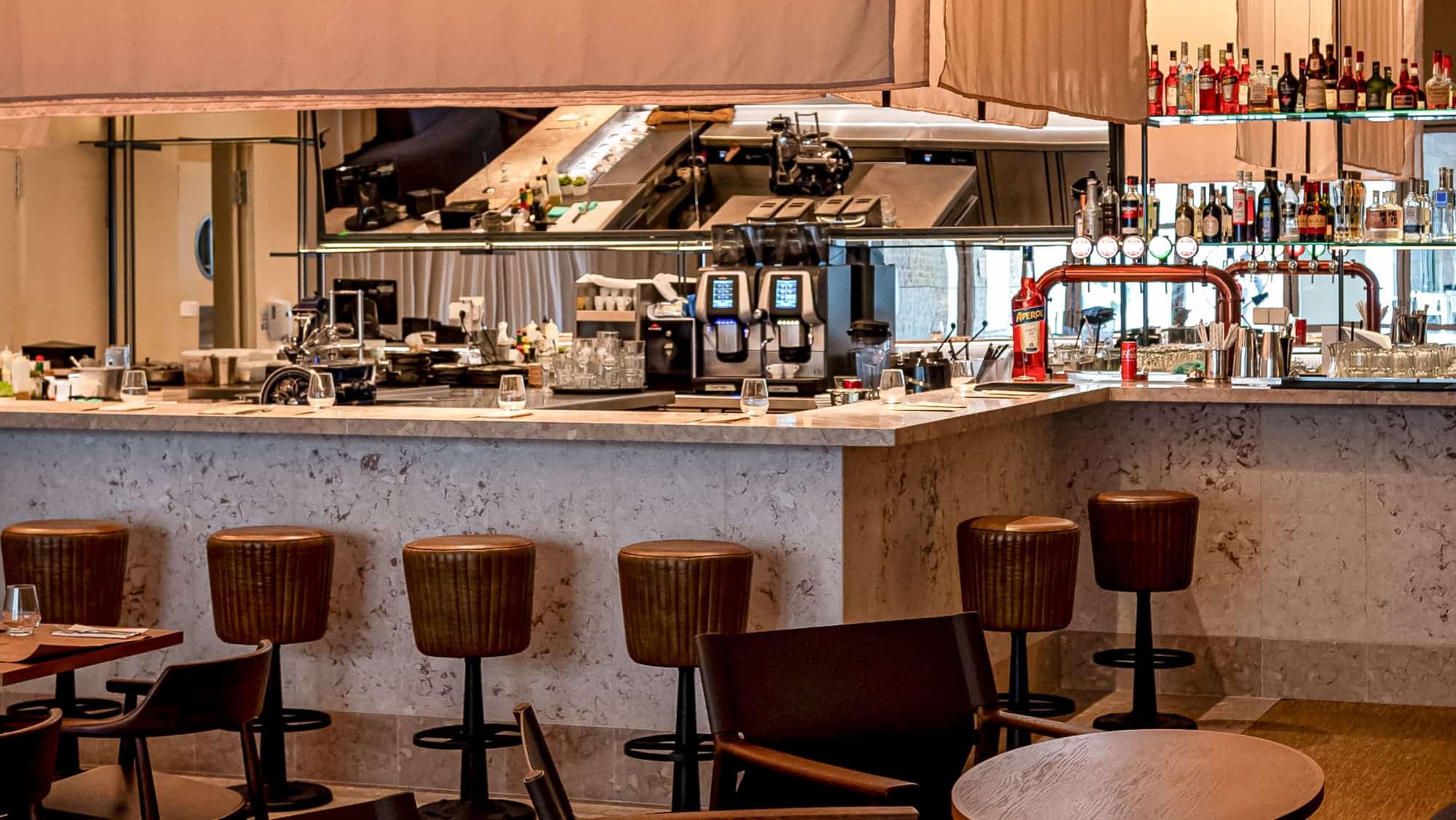 http://Imagem%20do%20bar%20do%20restaurante%20EsteOeste,%20moderno%20com%20bancada%20de%20mármore,%20várias%20banquetas%20de%20madeira%20ao%20redor,%20e%20uma%20área%20bem%20equipada%20com%20máquinas%20de%20café%20e%20bebidas%20alcoólicas%20ao%20fundo,%20ideal%20para%20um%20ambiente%20de%20bar%20e%20restaurante%20sofisticado.