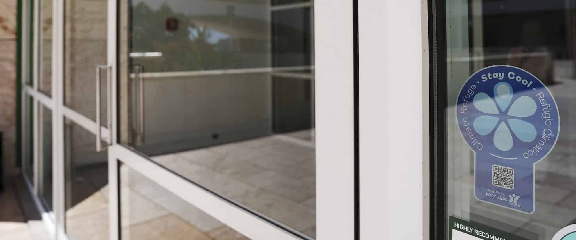 Porta de vidro de entrada com faixas brancas e adesivo azul no canto direito, exibindo texto de recomendação e um QR code, em ambiente interno com piso claro.
